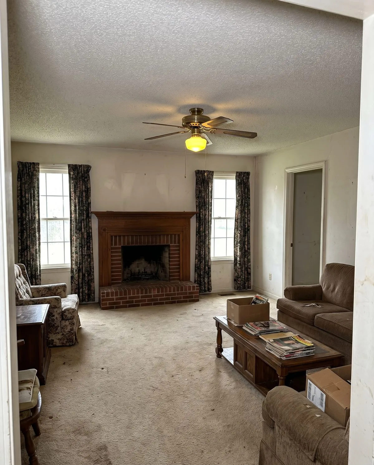 A dated living room before remodeling — brick fireplace, worn carpet, heavy curtains
